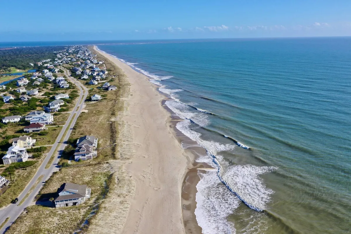 A wide stretch of sandy beach runs alongside the blue ocean, with a few charming coastal houses set back from the shore. The beach appears quiet and pristine, ideal for sunrise walks or spotting wildlife such as seabirds and, possibly, dolphins. The expansive sky and soft light enhance the feeling of peaceful immersion in the coastal landscape.