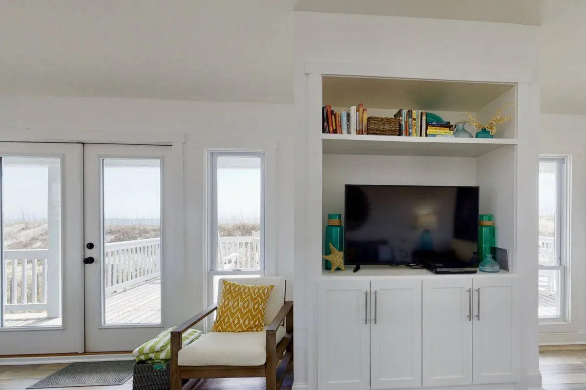 A comfortable living space featuring large windows overlooking the beach, a cozy seating area, and a television unit. The room is bathed in natural light, with furnishings arranged for relaxation and group activities.