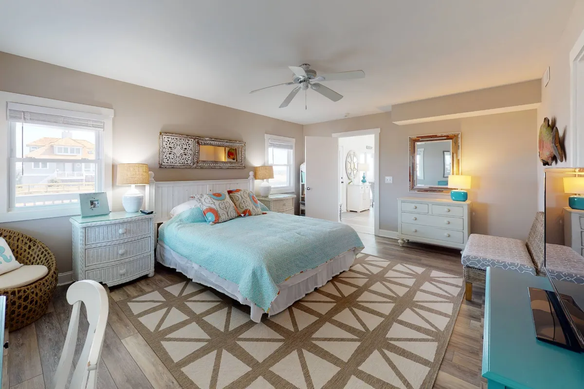 A tranquil bedroom with a comfortable bed adorned with decorative pillows, a patterned area rug, bedside lamps, and a decorative mirror. Gentle sunlight streams through the window, creating a calm and restful atmosphere ideal for relaxation.