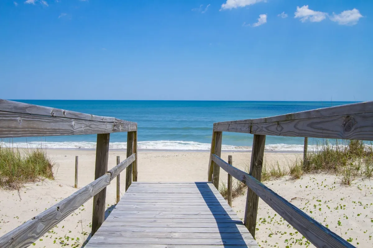 A wooden walkway leads across the dunes directly to a sandy beach and the ocean. The tranquil morning or afternoon light highlights the easy, private access to the shoreline, inviting families to step outside and start their day of beachfront adventure.