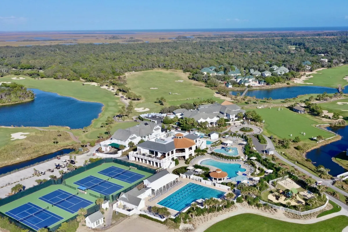 This image features the luxurious amenities of an exclusive island club, including elegant swimming pools, well-maintained tennis courts, a picturesque golf course, a bustling clubhouse, and serene water bodies. The scene is lively yet refined, capturing guests enjoying leisure activities against a backdrop of sunset hues.