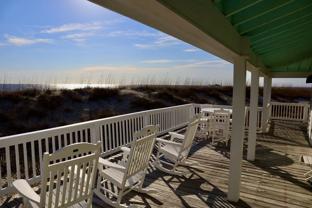 A peaceful scene from a wraparound deck or porch with classic rocking chairs overlooking dune grass and the sparkling ocean beyond. The sky is painted with soft sunset hues, inviting viewers to imagine relaxing while watching the evening settle in.