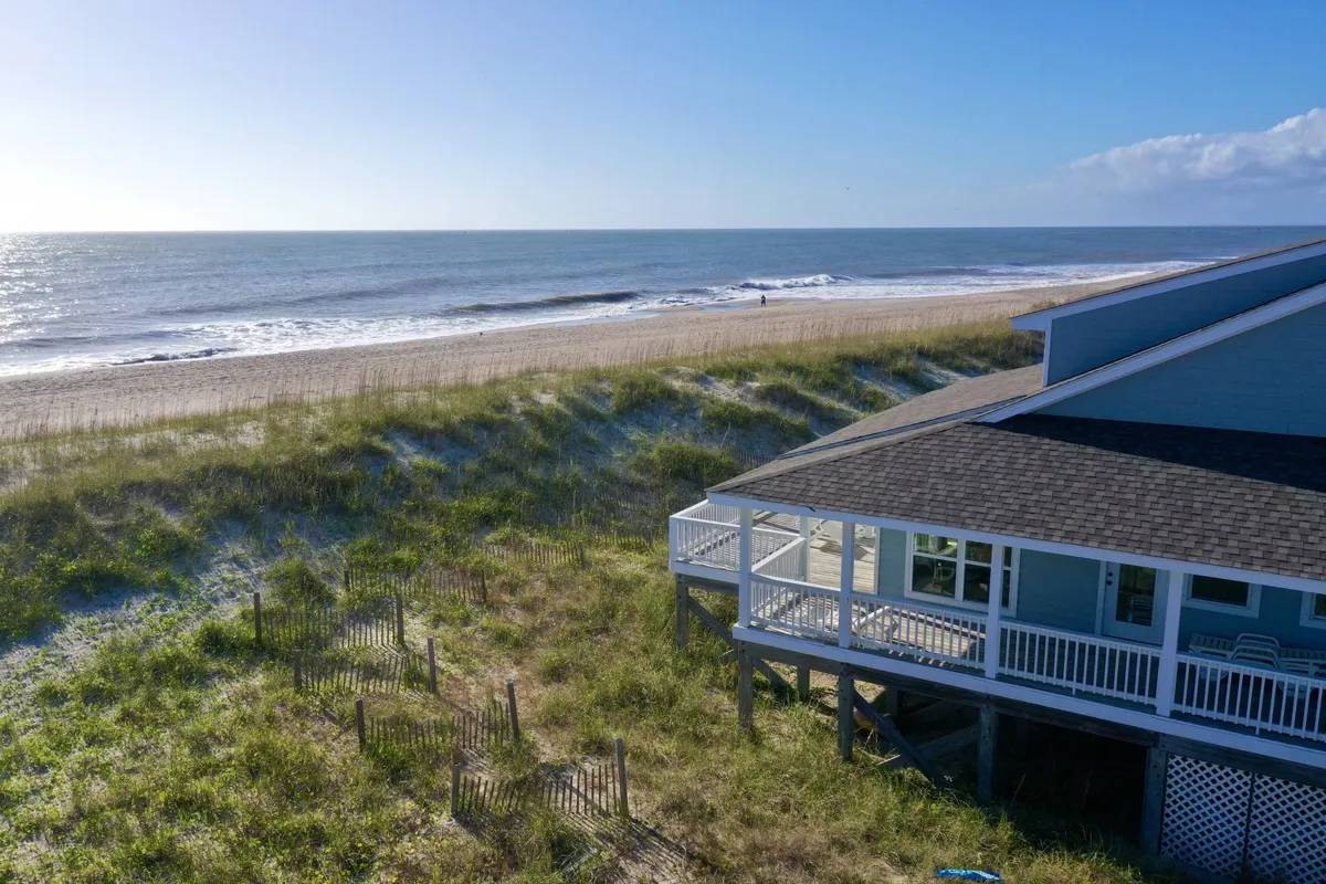 The image showcases an inviting beachfront home with a spacious wraparound deck overlooking rolling sand dunes and gentle ocean waves under a vibrant blue sky. The tranquil scene hints at both sunrise and sunset views, private beach access just steps away, and a serene natural setting ideal for relaxation and outdoor activities.