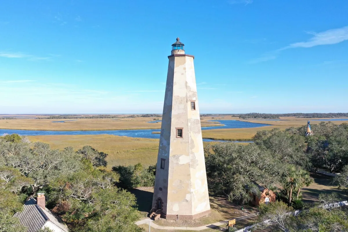 Old Baldy Lighthouse stands tall above the surrounding marshland, with lush greenery and a distant waterway visible. The historic structure is bathed in soft natural light, inviting exploration and offering a sense of Bald Head Island's unique character. The area is rich with birdlife and wildlife, making it ideal for adventure and history enthusiasts alike.