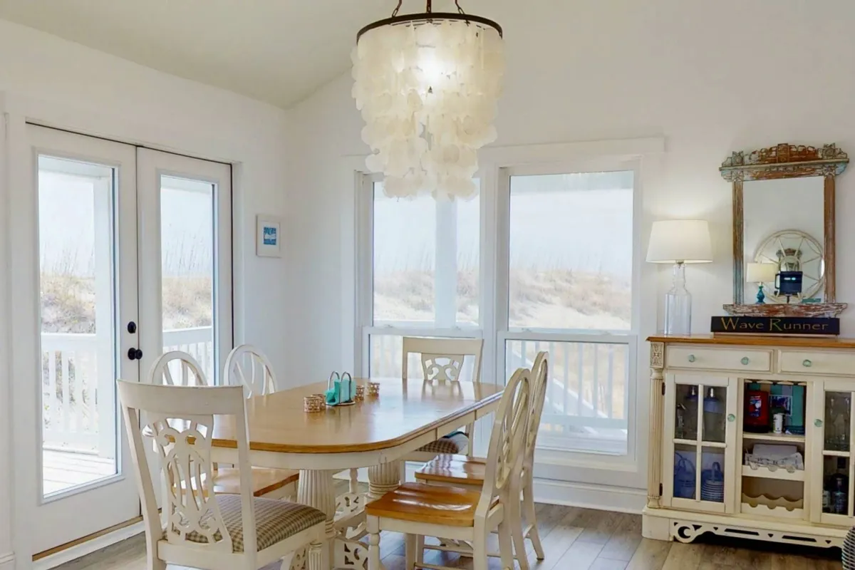 A bright dining area with a large table, elegant chandelier, decorative cabinet, and oversized windows that flood the space with natural light. The setup suggests lively meals and casual gatherings, with a view that hints at the outdoors beyond.