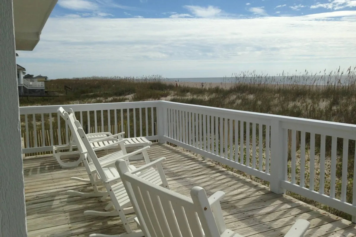 Two white rocking chairs are positioned on a porch overlooking rolling dunes and the vast ocean beyond. The soft light suggests either early morning or late afternoon, and the scene is framed by tall beach grasses, evoking a sense of seclusion and tranquility. The porch setting hints at peaceful moments spent in direct communion with nature.