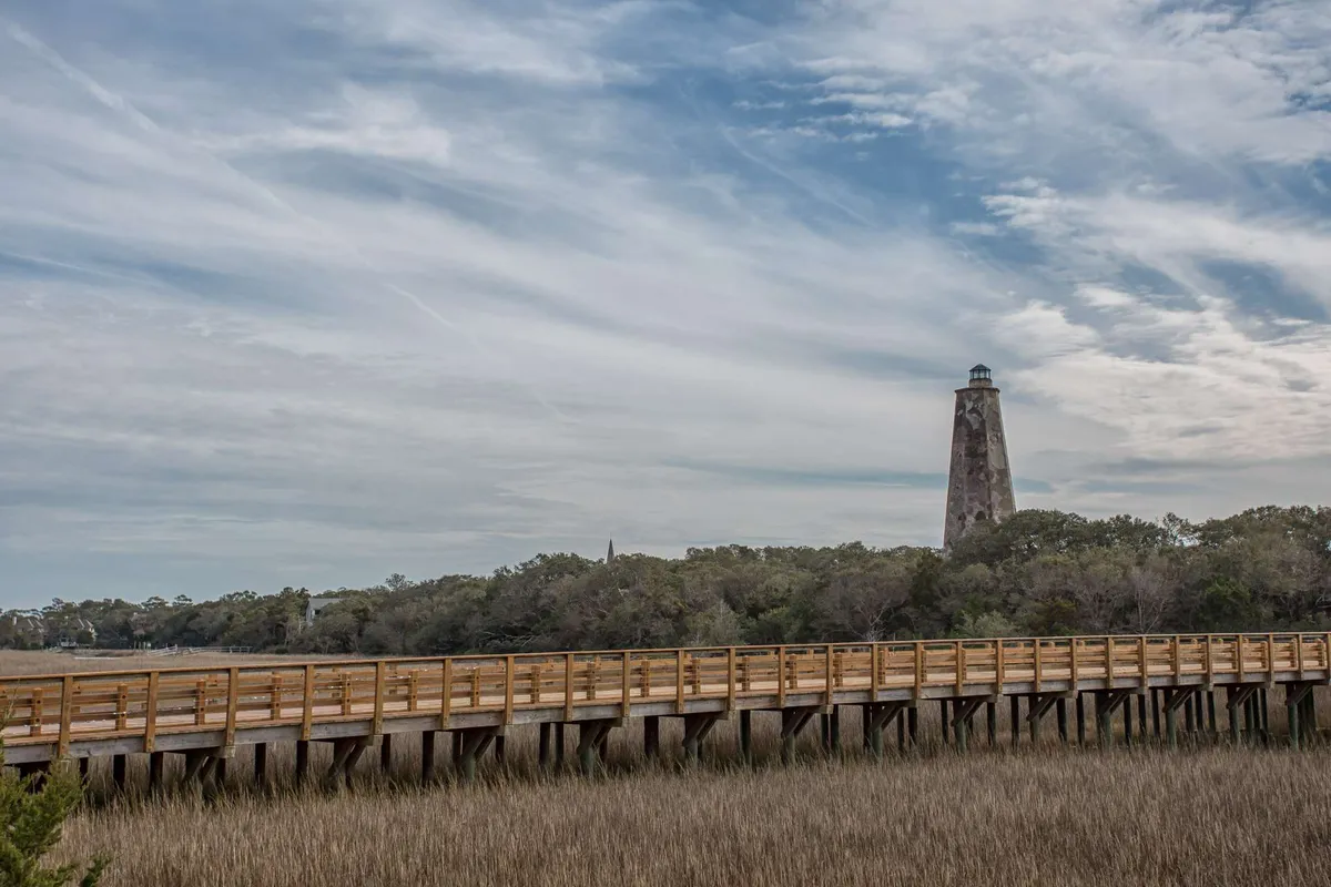 A picturesque view of the historic lighthouse with a wooden boardwalk winding through marsh grasses beneath a cloudy sky. The scene is tranquil yet iconic, evoking a sense of discovery and local heritage.