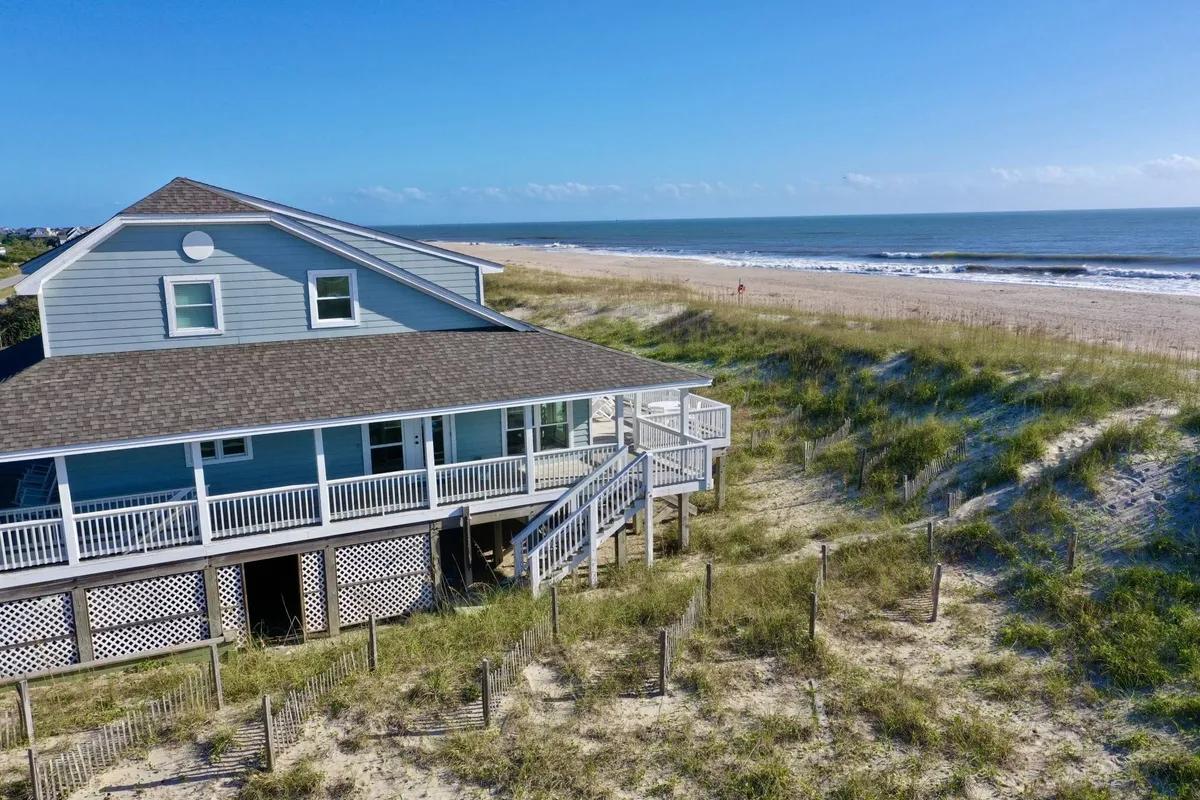 A beachfront property with a spacious wooden deck overlooking sand dunes and the ocean. The deck features ample seating, making it ideal for relaxing, dining, and enjoying the scenic views. The proximity to the beachfront is clear, with only a short path separating the deck from the sand.