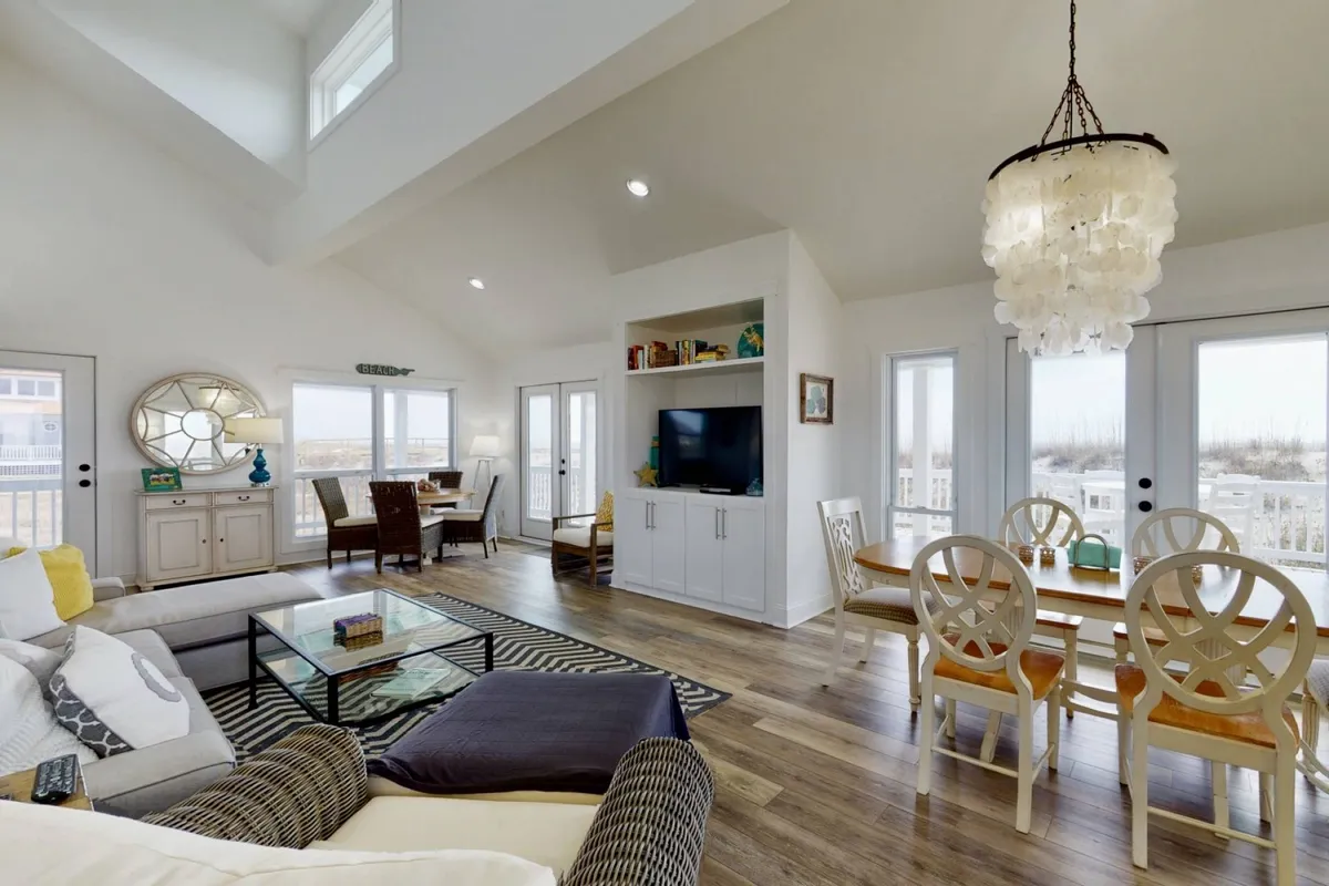 A spacious, cozy living room and dining area featuring comfortable sofas and a large window that frames expansive sunset views over the ocean. A chandelier hangs above the dining table, inviting communal meals, while the plush furnishings and soft lighting create a welcoming atmosphere.