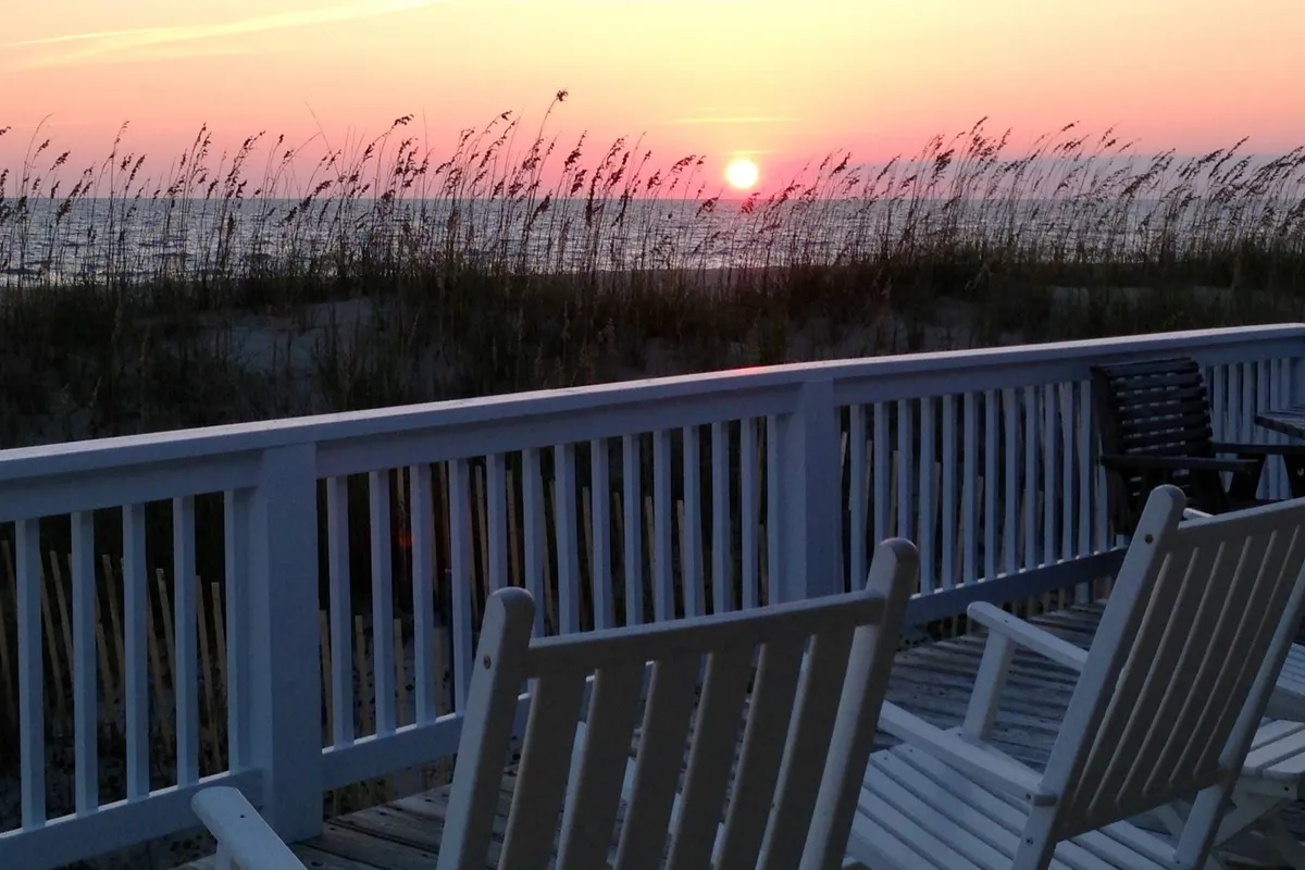 A serene beach scene at sunset, with chairs set up among soft beach grass facing the open ocean. The sun dips low on the horizon, casting a warm glow across the tranquil water, inviting relaxation and connection.