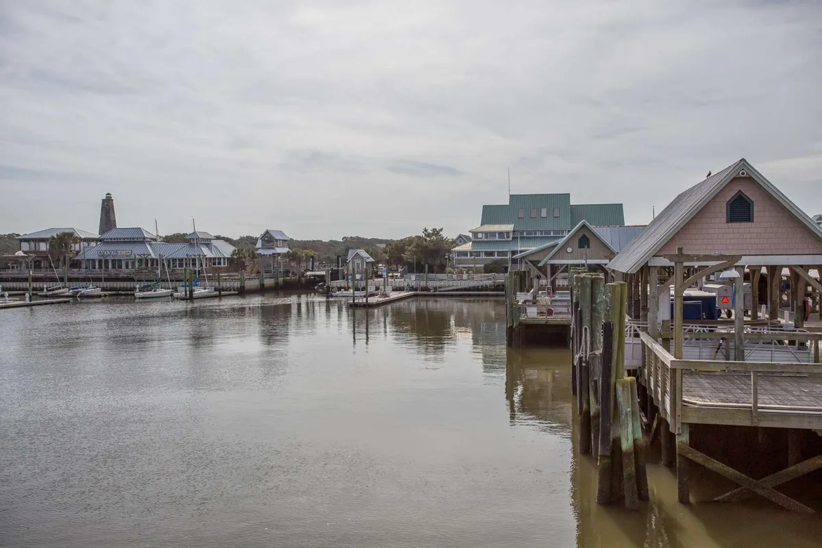 A lively marina scene with boats docked alongside waterfront buildings and a wooden pier. The setting suggests opportunities for boating, fishing, and exploring local cafes and boutiques nearby.