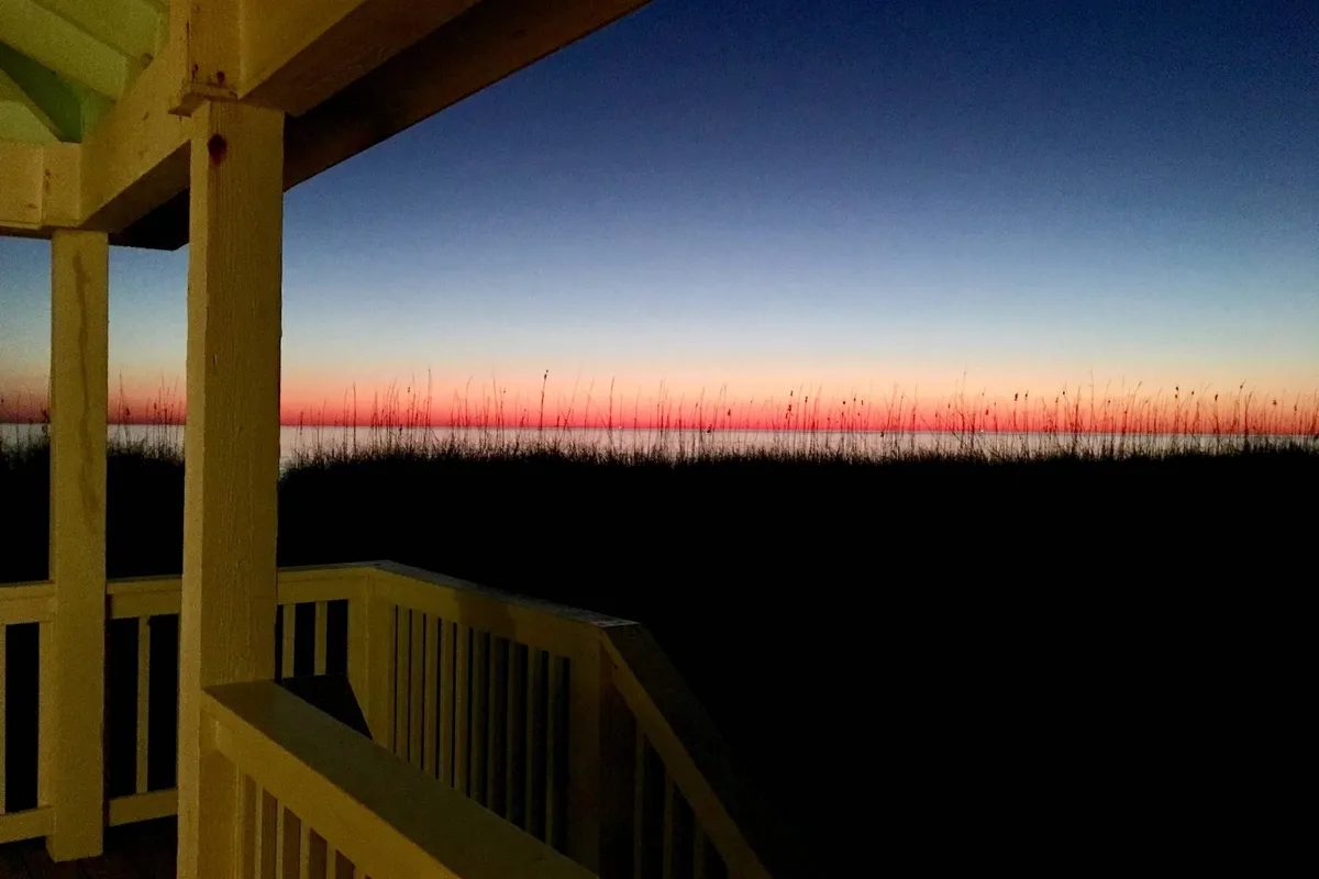 A serene sunset view from a porch overlooking the ocean, with silhouetted grasses adding depth and warmth. The inviting scene evokes moments spent gathering with loved ones, enjoying breathtaking natural beauty and the exclusivity of a private vantage point.