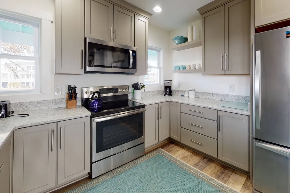 An inviting kitchen interior with stainless steel appliances, granite countertops, a colorful kitchen rug, and shelves adorned with decorative items. The kitchen appears ready for socializing over coffee, meal prep, or casual baking sessions with family and friends.